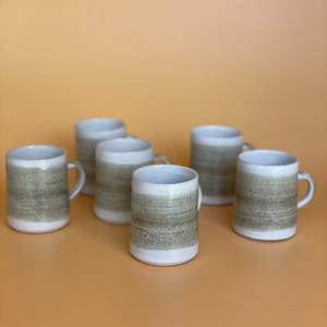 May include: A collection of six ceramic mugs. Each mug features a white base and rim, with a textured, earthy brown and green band around the middle. The mugs have a simple, curved handle and are arranged on an orange surface.