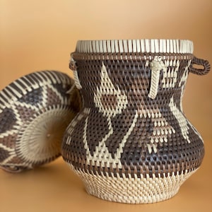 May include: A woven basket with a lid, featuring a brown and white geometric pattern. The basket has a handle and is made of natural materials.
