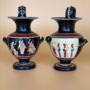 May include: Two black ceramic vases with handles and detailed artwork. The vases feature a black base, a red and white patterned band, and illustrations of figures in classical attire. The vases are displayed on a neutral background.