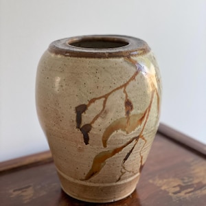 May include: A small, light brown ceramic vase with a brown and yellow floral design. The vase has a rounded body and a slightly flared lip.