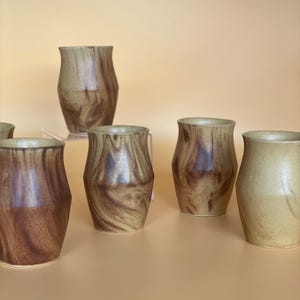 May include: Six handcrafted ceramic vases are displayed. The vases are cylindrical with a flared rim. The colour scheme features brown and tan tones, with a marbled pattern. The vases are arranged on a light orange surface.