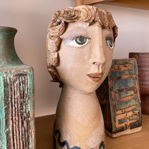 May include: A ceramic head sculpture with a tan finish, featuring sculpted brown hair, painted green eyes, and orange lips. The base of the neck is decorated with a black wavy line pattern. The sculpture has a smooth, matte texture throughout.