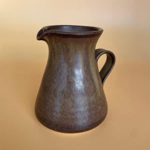 May include: A brown ceramic jug with a handle and a spout. The jug has a wide base that narrows towards the neck. The exterior has a mottled, earthy appearance, with variations in brown and tan hues. The handle is curved and attached to the side.