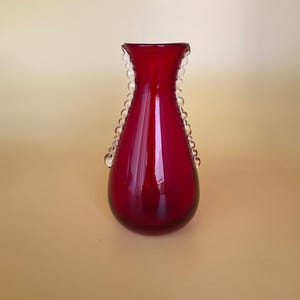 May include: A deep red glass vase with a rounded base and a scalloped rim. Two decorative glass loops are on the sides. The vase has a glossy finish. This decorative vase is suitable for flowers or as a decorative accent.