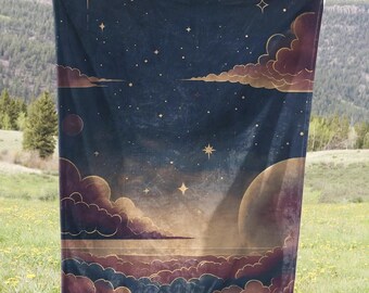 May include: A navy blue throw blanket with a celestial design featuring gold stars, clouds, and planets. The blanket is held up in front of a green grassy field with a mountain range in the background.