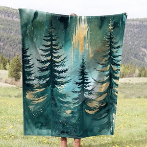 May include: A teal and gold throw blanket with a watercolor design of pine trees. The blanket is being held up in front of a grassy field with a mountain range in the background.