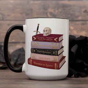 May include: White ceramic mug with a black handle and rim, featuring a stack of fantasy books with titles like "Touch Her and Die" and a skull. A small sword and skull sit atop the books.