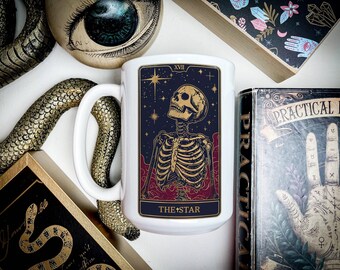 May include: White ceramic mug with a black and gold tarot card design. The card depicts a skeleton with outstretched arms against a dark blue background with stars. The card's title, "The Star", is printed in gold at the bottom.