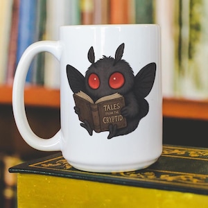 May include: White ceramic mug featuring a cartoon moth-like creature with red eyes, holding a book titled "TALES FROM THE CRYPTID." The creature is black with large wings. The mug has a large handle and is set against a blurred background of bookshelves.
