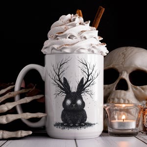 May include: White ceramic mug with a black and white illustration of a rabbit with antlers. The mug has whipped cream and cinnamon on top. The mug is sitting on a table with a skull and a candle.
