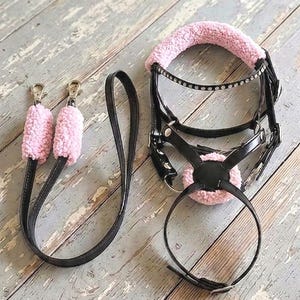 May include: A black leather horse bridle and matching reins featuring soft pink faux sheepskin padding on the crown piece, noseband, and rein grips. The bridle includes a browband adorned with a row of clear crystals and silver-toned metal hardware.