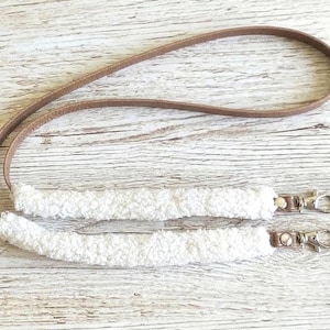 May include: A lanyard featuring a long, thin brown strap connected to two segments of white, textured, fuzzy material. Each end of the fuzzy sections is finished with a silver-toned metal swivel lobster clasp attached by a small brown tab.