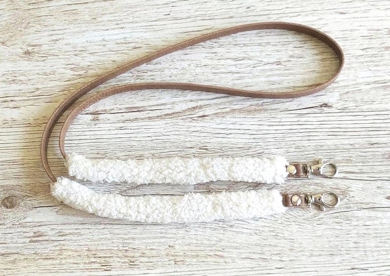 May include: A lanyard featuring a long, thin brown strap connected to two segments of white, textured, fuzzy material. Each end of the fuzzy sections is finished with a silver-toned metal swivel lobster clasp attached by a small brown tab.