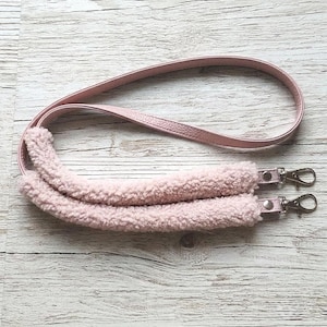 May include: A light pink bag strap featuring a smooth faux leather upper section and a plush, textured sherpa fleece shoulder pad. The strap includes two silver-toned metal swivel lobster clasps attached to the ends for secure bag attachment.