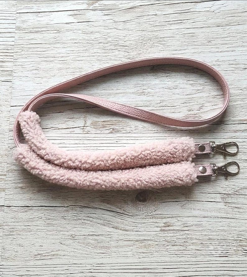 May include: A light pink bag strap featuring a smooth faux leather upper section and a plush, textured sherpa fleece shoulder pad. The strap includes two silver-toned metal swivel lobster clasps attached to the ends for secure bag attachment.