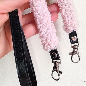 May include: A bag strap featuring a black faux leather base with two sections of textured, light pink curly faux shearling material. The strap includes silver-toned metal swivel lobster clasps attached to the ends with black leather tabs and silver rivets.