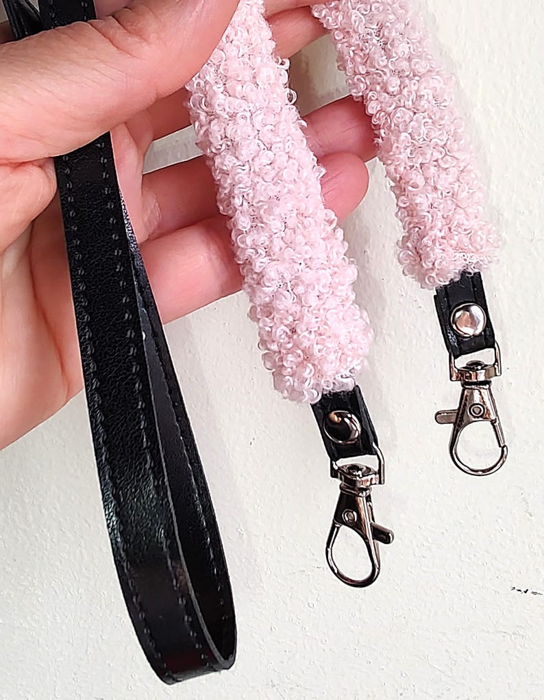 May include: A bag strap featuring a black faux leather base with two sections of textured, light pink curly faux shearling material. The strap includes silver-toned metal swivel lobster clasps attached to the ends with black leather tabs and silver rivets.