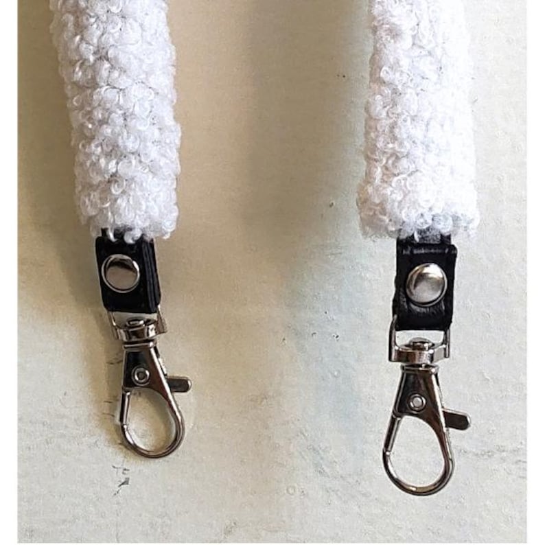 May include: A white textured sherpa strap featuring black leather tabs and silver-toned metal swivel lobster clasps. The strap has a thick, curly fleece-like surface texture and is secured to the metal hardware with silver-colored rivets.