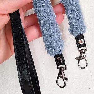 May include: A bag strap featuring a textured, light blue curly fleece material attached to black faux leather ends. The strap includes silver-toned metal swivel lobster clasps at both ends for secure attachment to a bag or accessory.