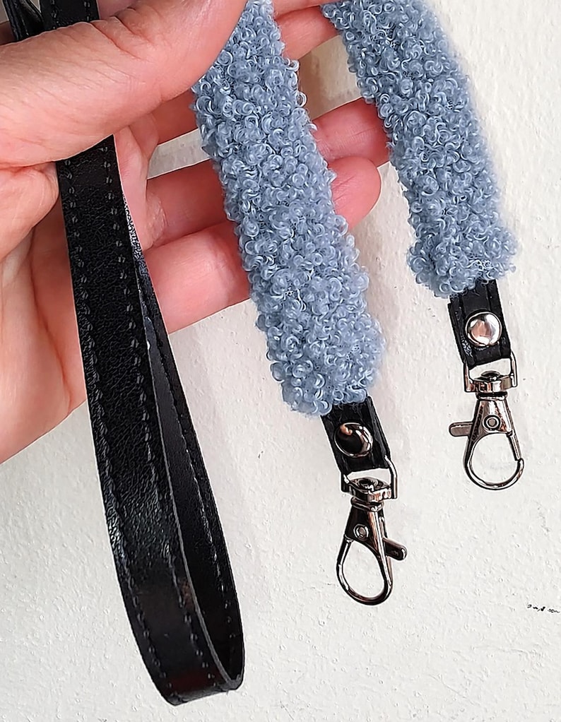 May include: A bag strap featuring a textured, light blue curly fleece material attached to black faux leather ends. The strap includes silver-toned metal swivel lobster clasps at both ends for secure attachment to a bag or accessory.