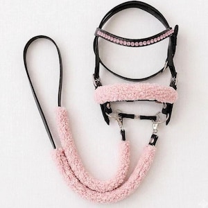 May include: A black horse bridle featuring a browband adorned with a row of pink crystals. The noseband and the lower sections of the reins are covered in soft, light pink faux sheepskin padding. The bridle includes silver-toned metal hardware.