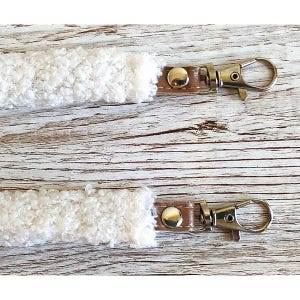 May include: Two white textured sherpa fabric straps, each featuring a brown leather-like tab secured with a silver-tone metal rivet and a matching silver-tone swivel lobster clasp hook attached to the end of each strap for secure fastening.