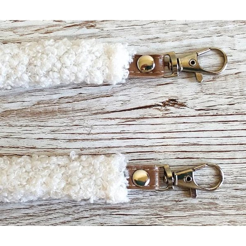 May include: Two white textured sherpa fabric straps, each featuring a brown leather-like tab secured with a silver-tone metal rivet and a matching silver-tone swivel lobster clasp hook attached to the end of each strap for secure fastening.