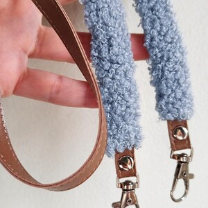 May include: A bag strap featuring two sections of light blue textured sherpa-style fabric attached to brown faux leather ends. Each end is secured with a silver-toned metal snap button and includes a silver-toned metal swivel lobster clasp hook.