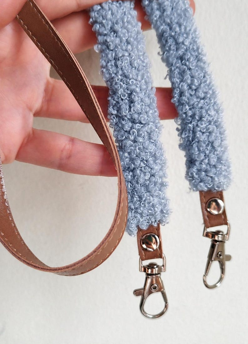 May include: A bag strap featuring two sections of light blue textured sherpa-style fabric attached to brown faux leather ends. Each end is secured with a silver-toned metal snap button and includes a silver-toned metal swivel lobster clasp hook.