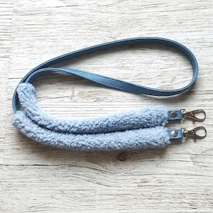 May include: A light blue bag strap featuring a smooth faux leather upper section and a plush, fuzzy sherpa-style fleece shoulder pad. The strap includes silver-toned metal swivel lobster claw clasps at both ends for secure attachment to a bag.
