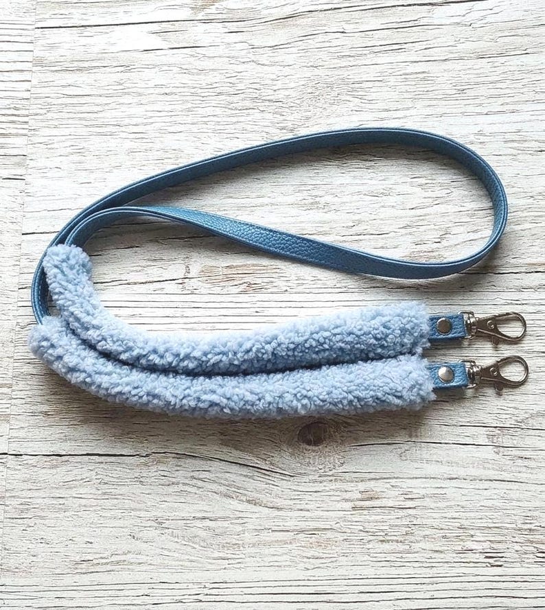 May include: A light blue bag strap featuring a smooth faux leather upper section and a plush, fuzzy sherpa-style fleece shoulder pad. The strap includes silver-toned metal swivel lobster claw clasps at both ends for secure attachment to a bag.