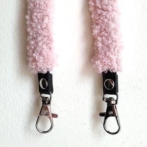 May include: Two light pink, fuzzy, textured fabric straps featuring black leather-like tabs secured with silver-toned metal snaps. Each strap terminates in a polished silver-toned metal swivel lobster clasp for attachment to a bag or accessory.