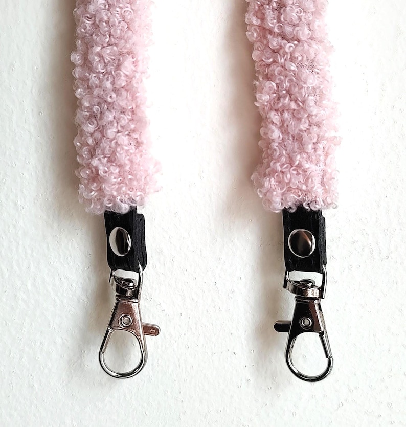 May include: Two light pink, fuzzy, textured fabric straps featuring black leather-like tabs secured with silver-toned metal snaps. Each strap terminates in a polished silver-toned metal swivel lobster clasp for attachment to a bag or accessory.