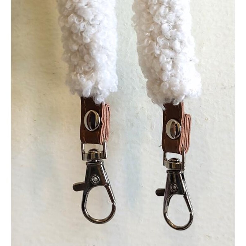 May include: Two white sherpa-style textured fabric straps featuring tan faux leather tabs secured with silver-tone metal snaps. Each strap end is finished with a silver-tone metal swivel lobster clasp for attachment, providing a functional design.