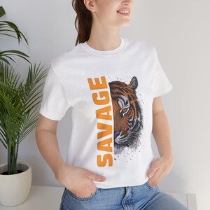 Puede incluir: Camiseta blanca con la cara de un tigre y la palabra "SAVAGE" en naranja. La cara del tigre es marrón, negra y blanca, con una expresión feroz. La camiseta es de estilo cuello redondo clásico.