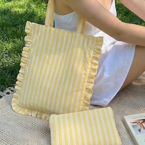 Op de afbeelding: Een geel en wit gestreepte tote bag met gerimpelde randen en een bijpassend klein etui. De tas heeft een rechthoekige vorm met verticale strepen en een eenvoudige handgreep. Het etui is rechthoekig en heeft een ritssluiting.