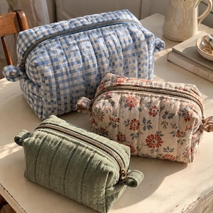 Puede incluir: Tres estuches acolchados rectangulares con cremalleras metálicas y pestañas laterales. Un estuche presenta un patrón de cuadros vichy azul y blanco, otro un estampado floral con flores rojas y azules sobre fondo crema, y el tercero es verde salvia.