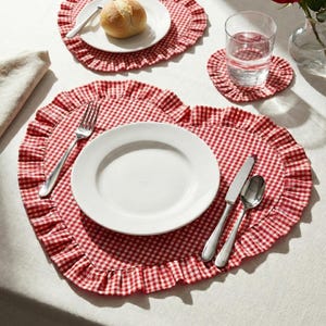 Op de afbeelding: Een tafelsetting met hartvormige placemats met een rood-wit ruitpatroon en gerimpelde randen. Witte borden, bestek, een broodje en een glas water zijn op de placemats geplaatst. Een rode roos in een vaas is ook aanwezig.