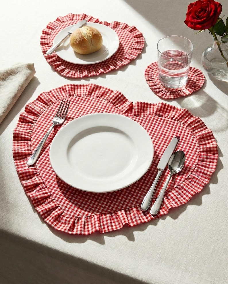 Puede incluir: Una mesa puesta con manteles individuales en forma de coraz&oacute;n con un patr&oacute;n de cuadros rojos y blancos y bordes con volantes. Platos blancos, cubiertos, un panecillo y un vaso de agua est&aacute;n colocados sobre los manteles. Tambi&eacute;n hay una rosa roja en un jarr&oacute;n.