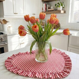 Puede incluir: Un ramo de tulipanes naranjas y amarillos en un jarr&oacute;n de cristal transparente sobre un mantel individual de cuadros rojos y blancos con borde con volantes. La cocina tiene armarios blancos.