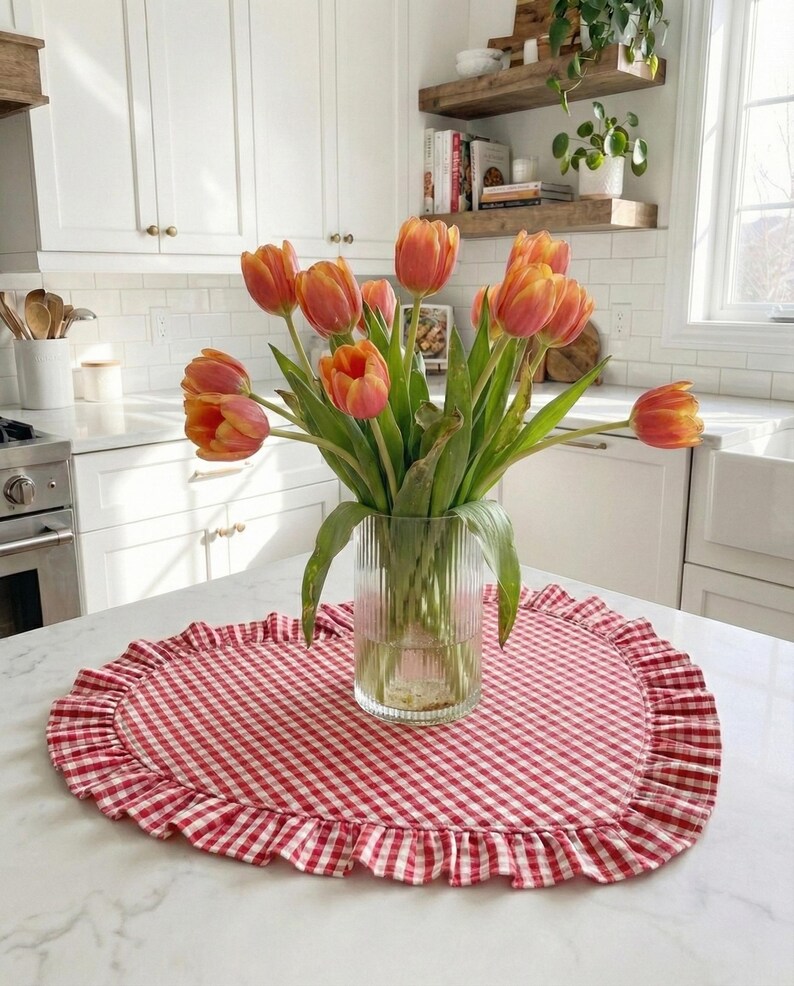 Puede incluir: Un ramo de tulipanes naranjas y amarillos en un jarr&oacute;n de cristal transparente sobre un mantel individual de cuadros rojos y blancos con borde con volantes. La cocina tiene armarios blancos.