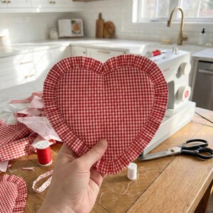 Puede incluir: Un objeto en forma de coraz&oacute;n a cuadros rojos y blancos con un borde con volantes se sostiene en una mano. Suministros de costura, incluyendo carretes de hilo y tijeras, est&aacute;n sobre una mesa de madera. Una m&aacute;quina de coser est&aacute; en el fondo.