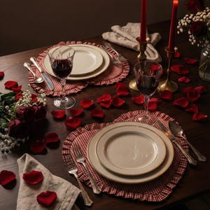 Puede incluir: Una mesa rom&aacute;ntica con dos cubiertos. Cada uno incluye un plato blanco con borde dorado, cubiertos y una copa de vino llena de vino tinto. Manteles individuales rojos en forma de coraz&oacute;n, p&eacute;talos de rosa y velas rojas encendidas completan la escena.