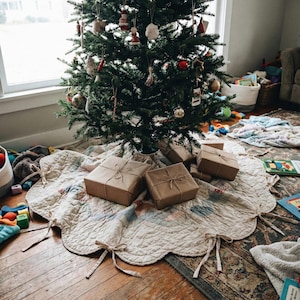 May include: A cream-colored quilted tree skirt with scalloped edges and fabric ties, positioned under a decorated Christmas tree. Several rectangular gift boxes wrapped in plain brown paper and tied with twine sit on the fabric skirt.