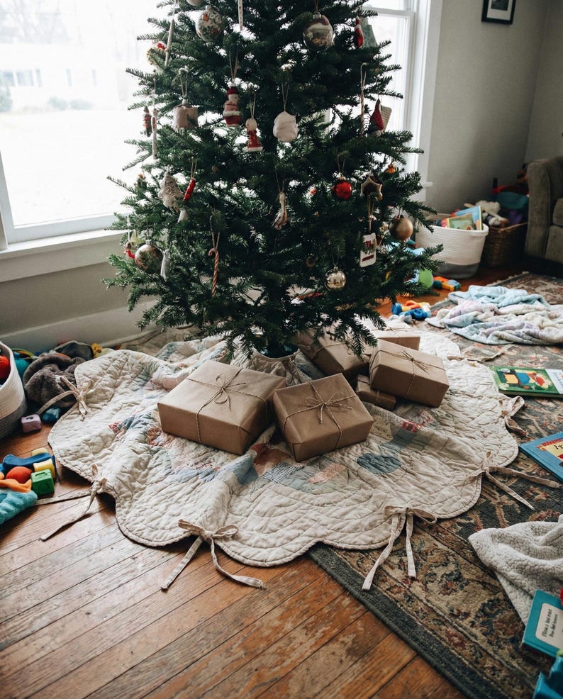 May include: A cream-colored quilted tree skirt with scalloped edges and fabric ties, positioned under a decorated Christmas tree. Several rectangular gift boxes wrapped in plain brown paper and tied with twine sit on the fabric skirt.