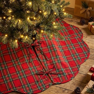 May include: A round red and green plaid fabric tree skirt with scalloped edges and two sets of fabric ties for closure. The tree skirt is positioned underneath a Christmas tree with warm white lights and is made of a woven textile material.