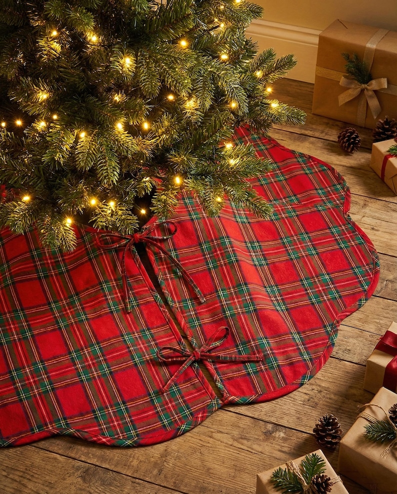 May include: A round red and green plaid fabric tree skirt with scalloped edges and two sets of fabric ties for closure. The tree skirt is positioned underneath a Christmas tree with warm white lights and is made of a woven textile material.