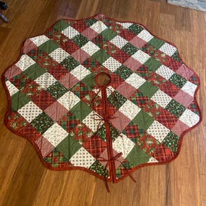 May include: A round, quilted Christmas tree skirt featuring a patchwork design of red, green, and white fabric squares. The fabric patterns include stripes, plaid, and floral motifs. It has a scalloped edge and three fabric ties for closure.