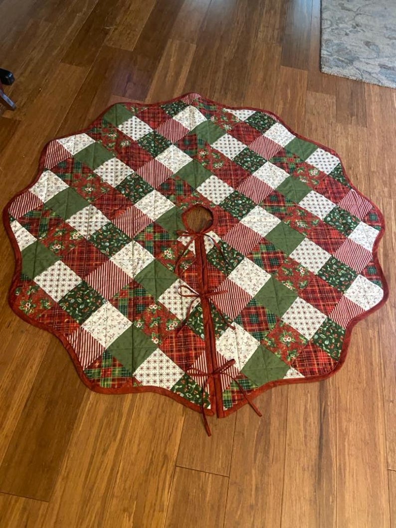 May include: A round, quilted Christmas tree skirt featuring a patchwork design of red, green, and white fabric squares. The fabric patterns include stripes, plaid, and floral motifs. It has a scalloped edge and three fabric ties for closure.
