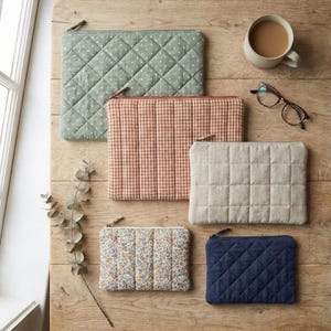 Puede incluir: Vista aérea de cinco estuches acolchados en varios colores y patrones, incluyendo verde salvia con lunares blancos, cuadros de melocotón, crema, floral y azul marino. Una taza de café y gafas están sobre una mesa de madera.